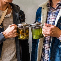 Two men smiling while holding up mugs of tea, celebrating a moment together.