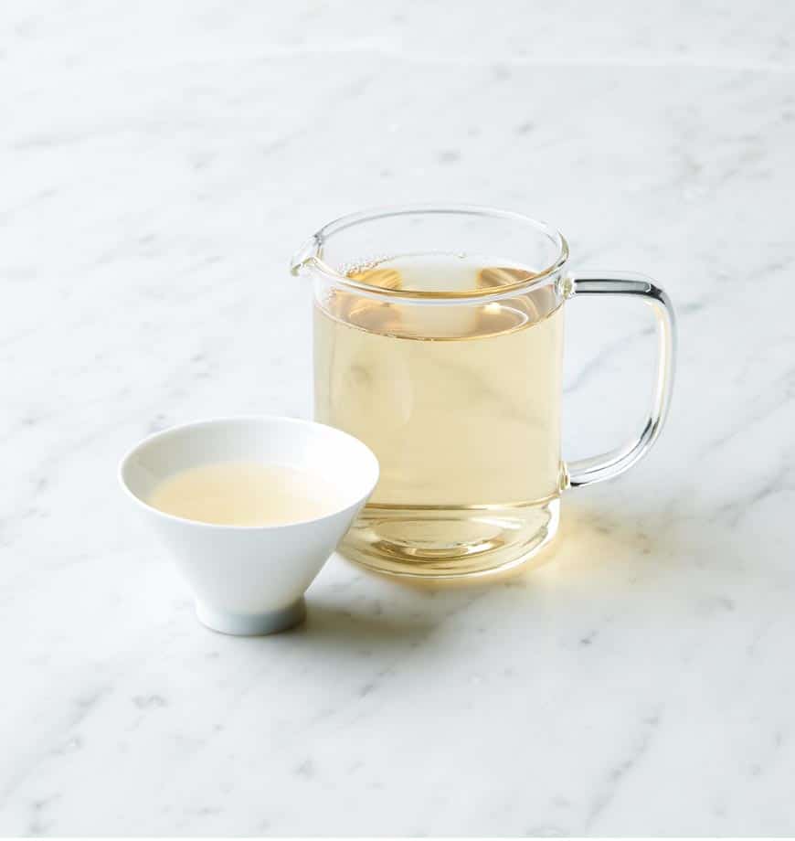 A pitcher of green tea alongside a cup, labeled as Downy Sprout white tea, set on a wooden table.