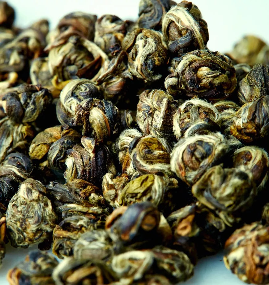 A pile of dried jasmine pearl green tea leaves on a white surface, showcasing their delicate, rolled appearance.