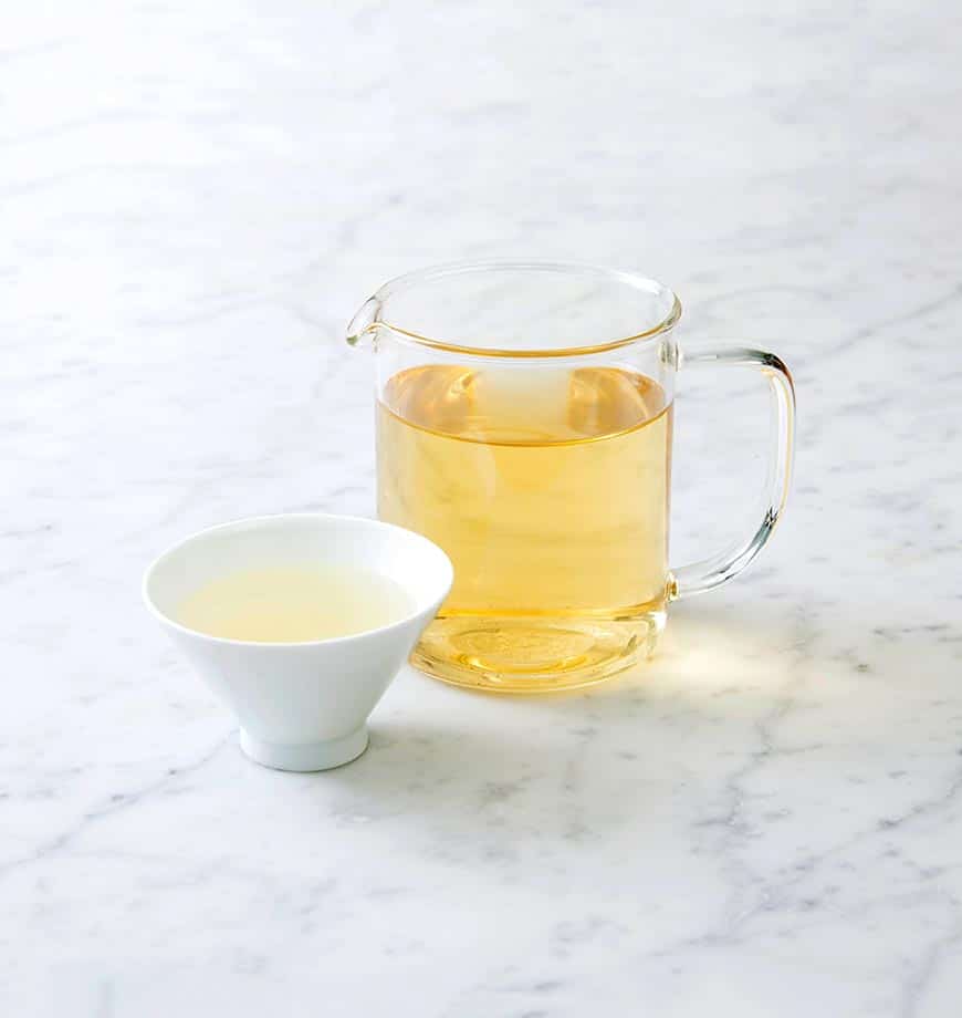 A glass pitcher of Dandelion Detox herbal infusion with a cup of tea beside it on a wooden table.