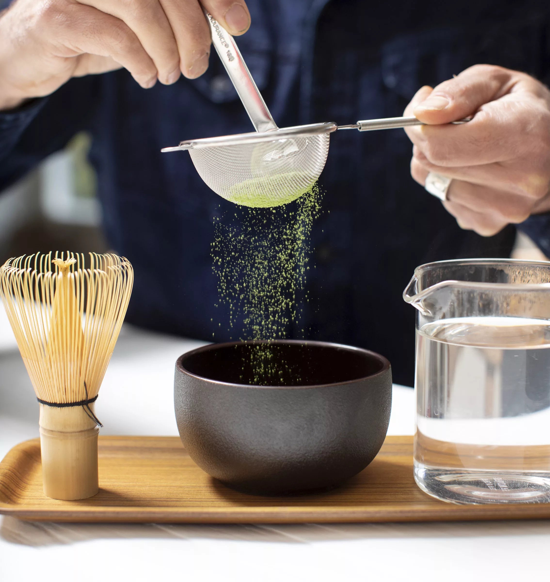 A hand pours green tea into a black matcha bowl, showcasing the tea's vibrant color.