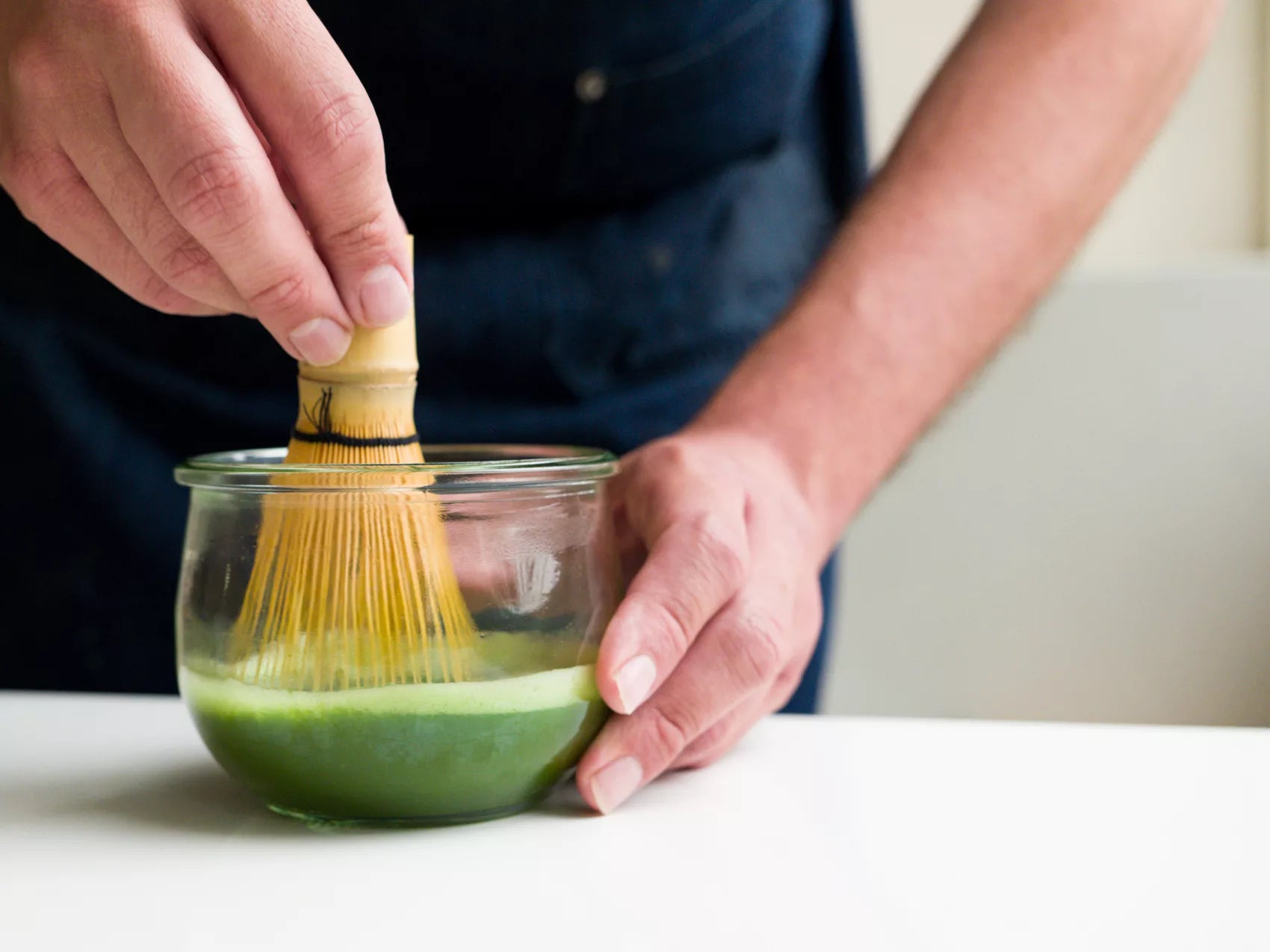 A person uses a brush to mix organic Mainichi Matcha green tea, resulting in a rich green liquid in a bowl.