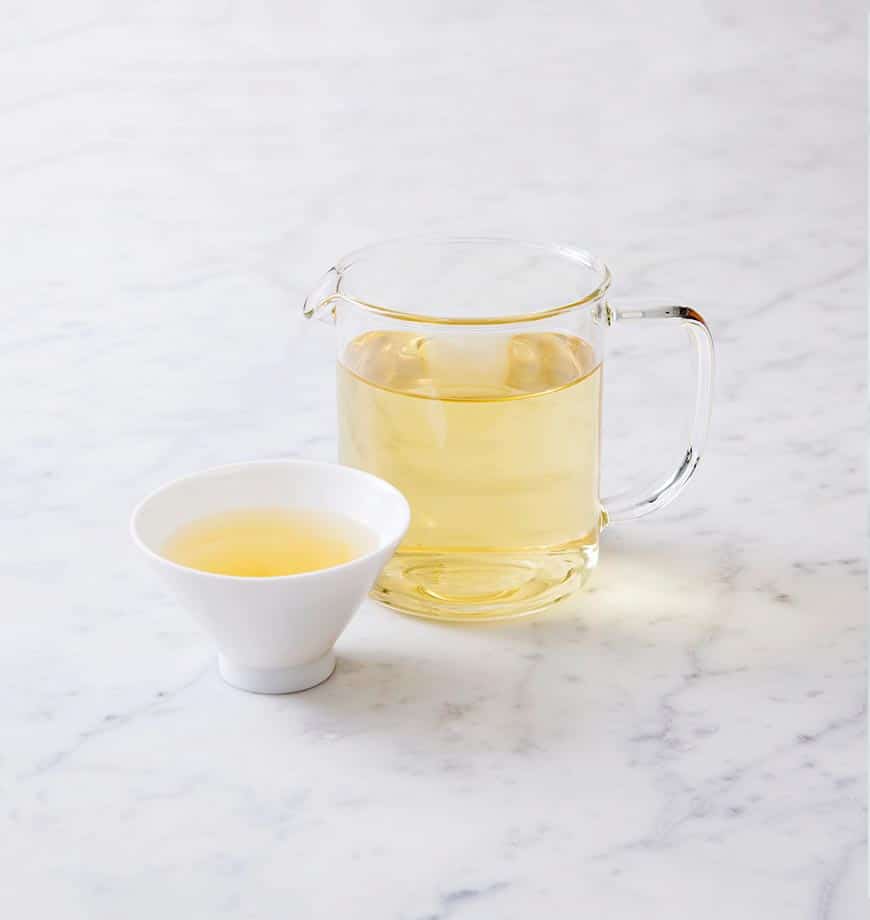 A pitcher of green tea alongside a cup filled with chamomile blossom herbal infusion.
